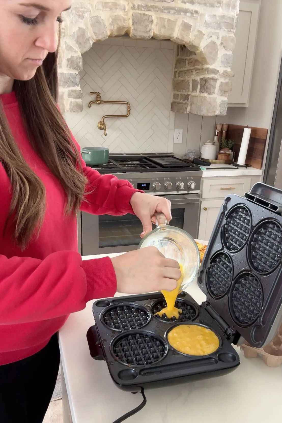 A woman in a red sweater pours a whisked egg mixture into a waffle maker to prepare an egg patty for a breakfast sandwich, standing in a cozy kitchen with a rustic stone oven hood in the background.